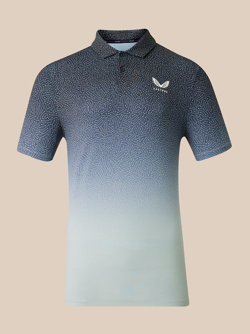 A short-sleeve polo shirt with a dark blue and light blue gradient pattern, featuring a small white wing logo on the left chest that reads 'CASTORE. The shirt is displayed on a solid beige background.