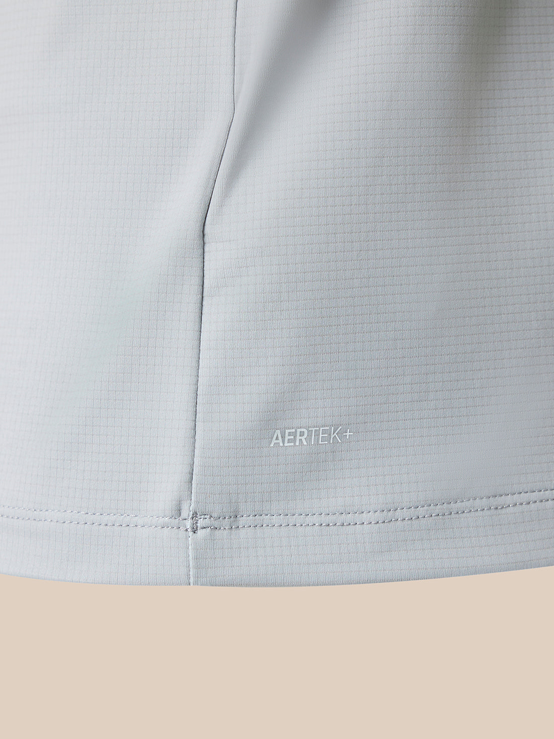 Close-up of a light gray athletic fabric with a fine grid texture, showing a vertical seam and the AERTEK+ logo printed subtly near the lower front. The fabric is shown against a solid beige background.