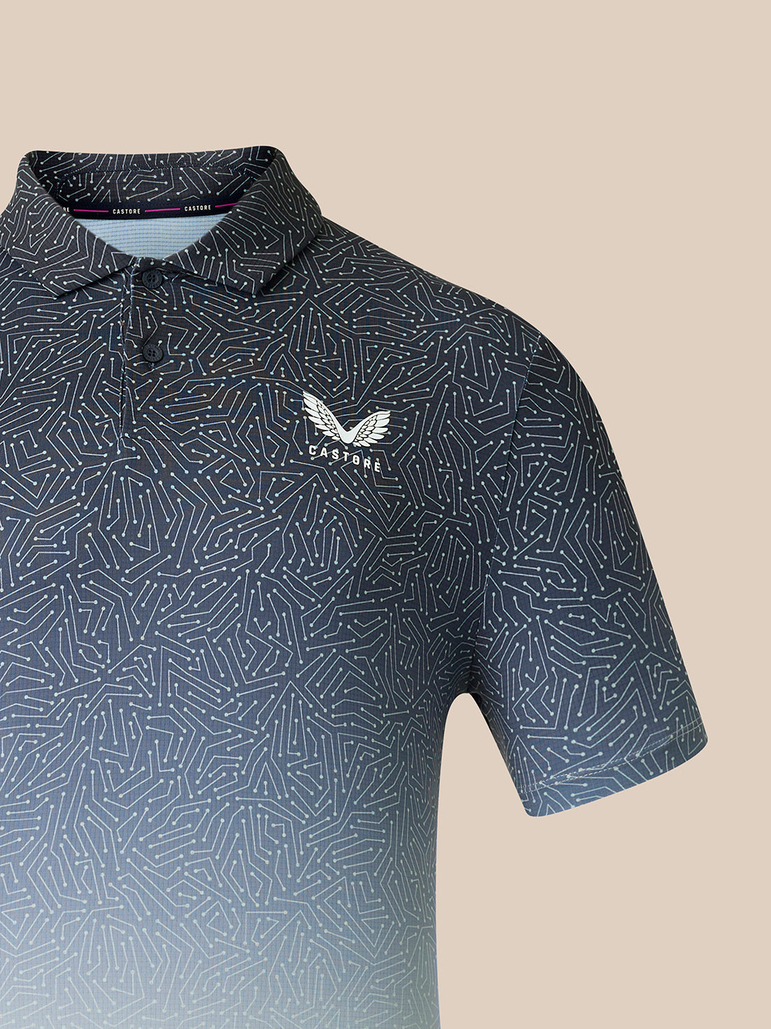 A close-up of a short-sleeve polo shirt with a dark blue and gray geometric pattern, featuring a white embroidered Castore logo on the left chest, set against a solid beige background.