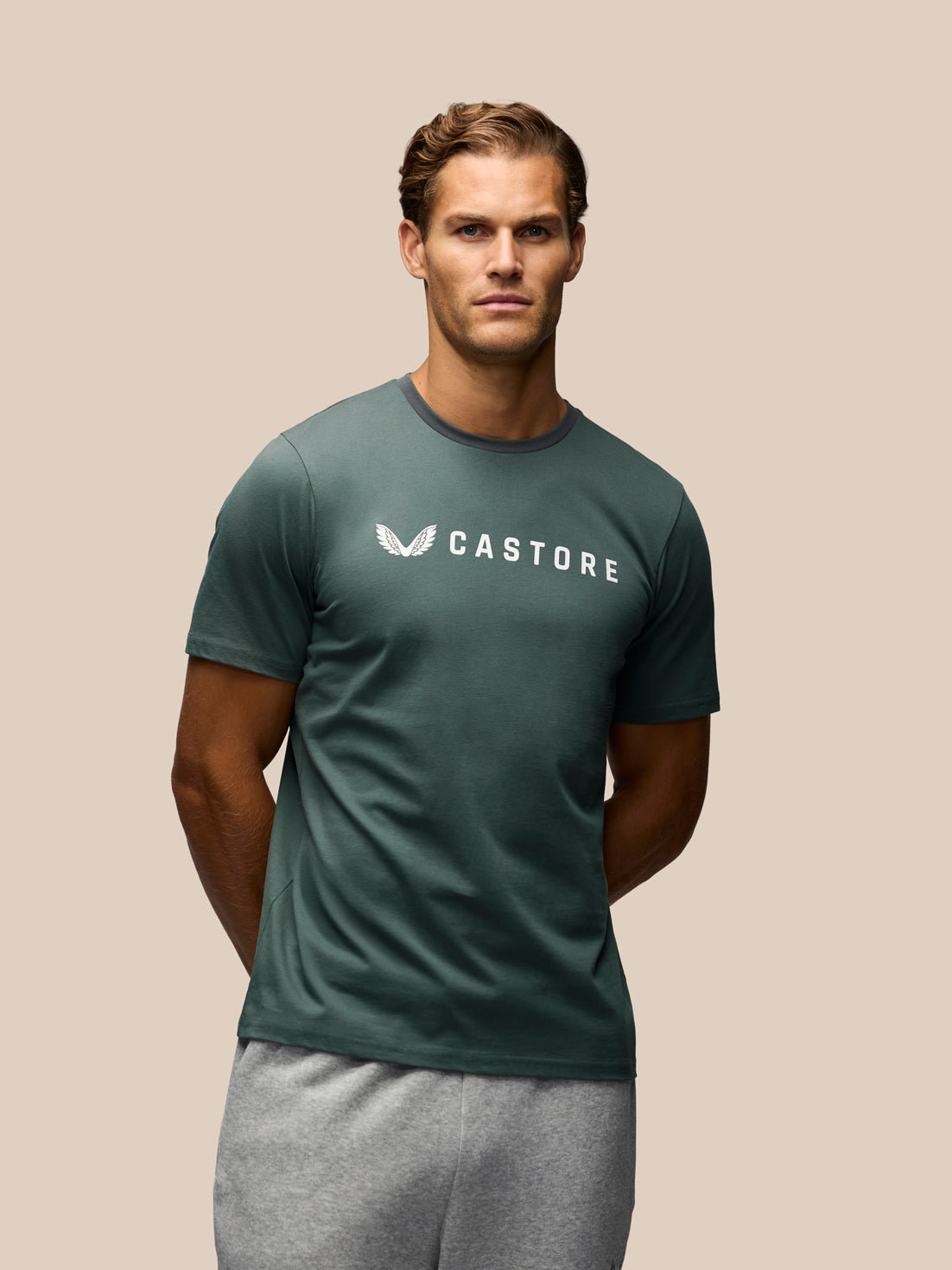 A man wearing a dark green Castore short-sleeve t-shirt with a white logo on the chest, paired with gray sweatpants, set against a solid beige background.