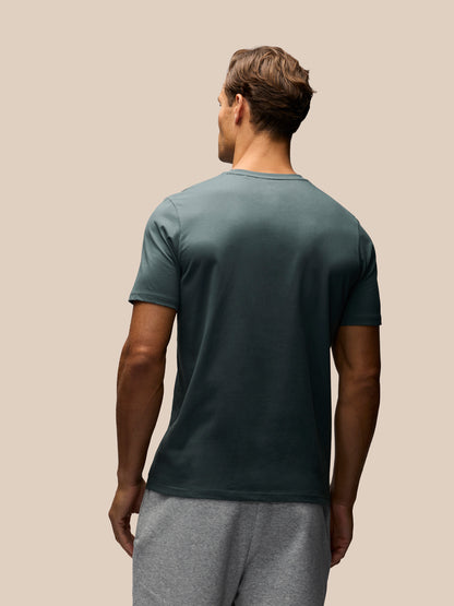 A man viewed from behind wearing a dark teal-green short-sleeve t-shirt and gray sweatpants, standing against a solid beige background.