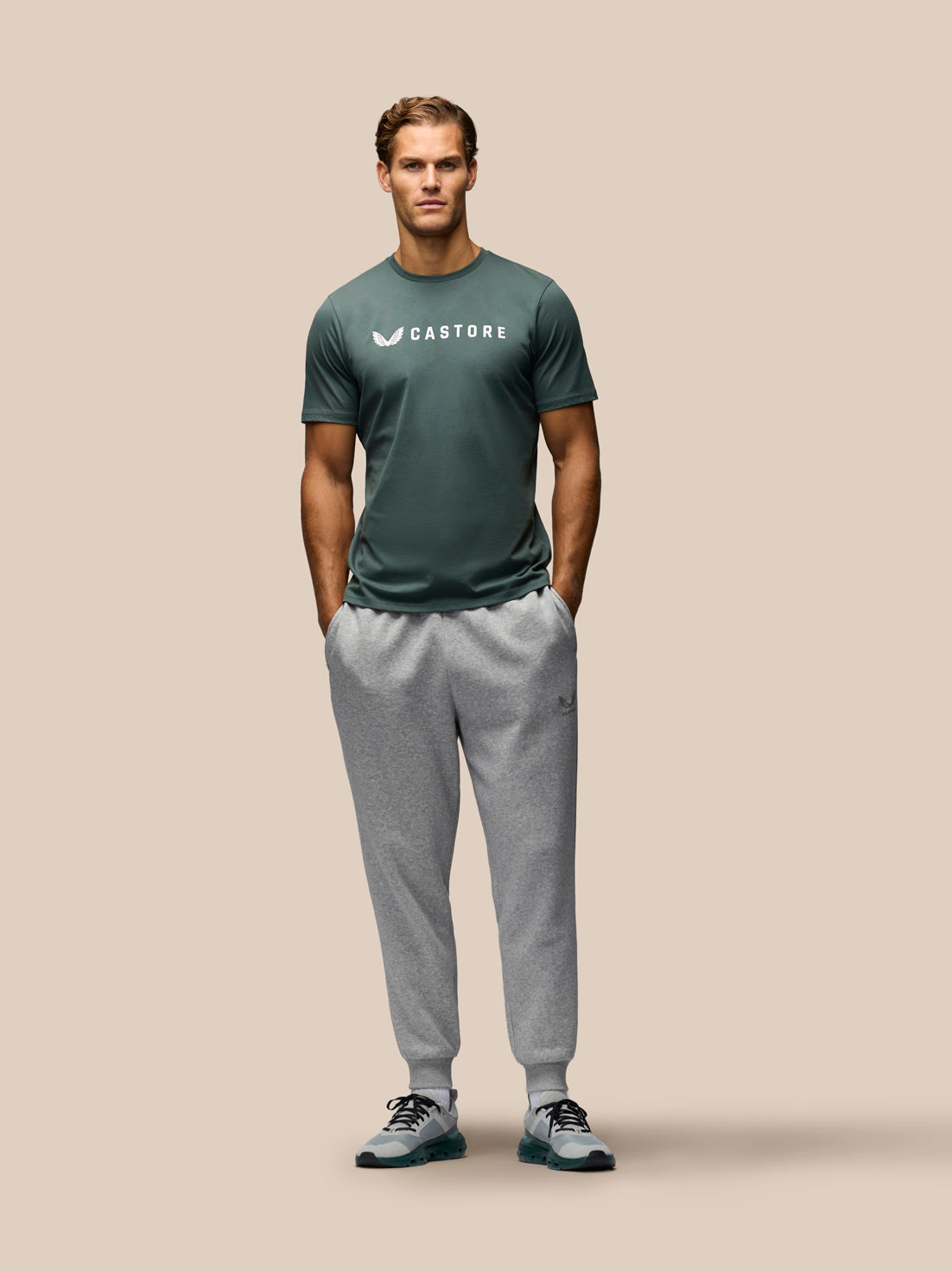 A man stands against a solid beige background wearing a dark green Castore athletic t-shirt and light gray jogger sweatpants.