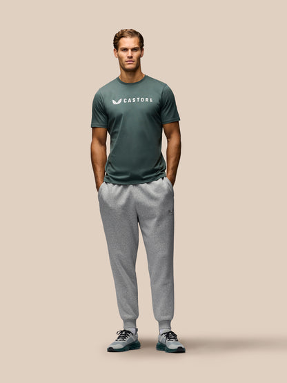 A man stands against a solid beige background wearing a dark green Castore athletic t-shirt and light gray jogger sweatpants.