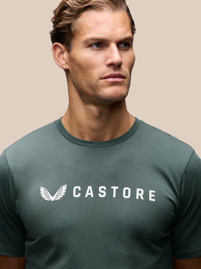 A man wearing a dark green Castore t-shirt with a white wing logo and brand name printed on the chest, set against a solid beige background.