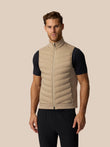 A man wearing a beige puffer vest over a black short-sleeve athletic shirt and black pants, standing against a solid beige background.