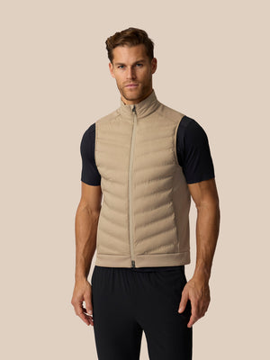 A man wearing a beige puffer vest over a black short-sleeve athletic shirt and black pants, standing against a solid beige background.