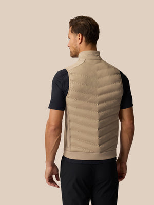 A man shown from the back wearing a beige puffer vest over a black short-sleeve shirt and black pants, against a solid light beige background.