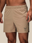 Beige athletic shorts worn by a model, featuring an elastic waistband and a dark gray wing logo with the text 'CASTORE REISS' on the lower left leg.
