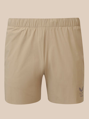 Beige athletic shorts with an elastic waistband and a small dark logo on the lower left leg, shown against a solid beige background.