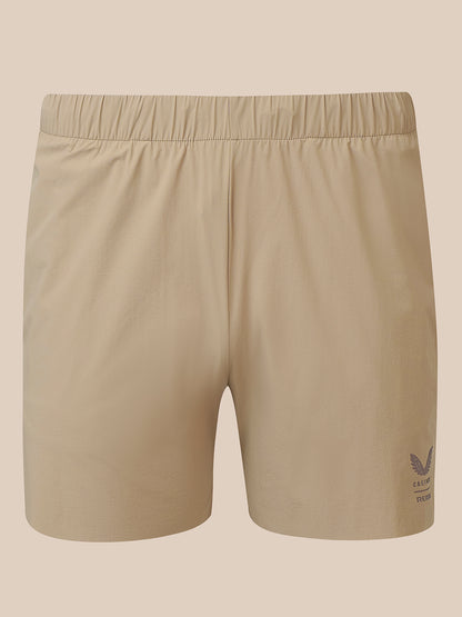 Beige athletic shorts with an elastic waistband and a small dark logo on the lower left leg, shown against a solid beige background.