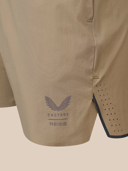 Close-up of beige Castore Reiss athletic shorts showing the side seam, perforated side panel, and a dark gray wing logo with brand text.