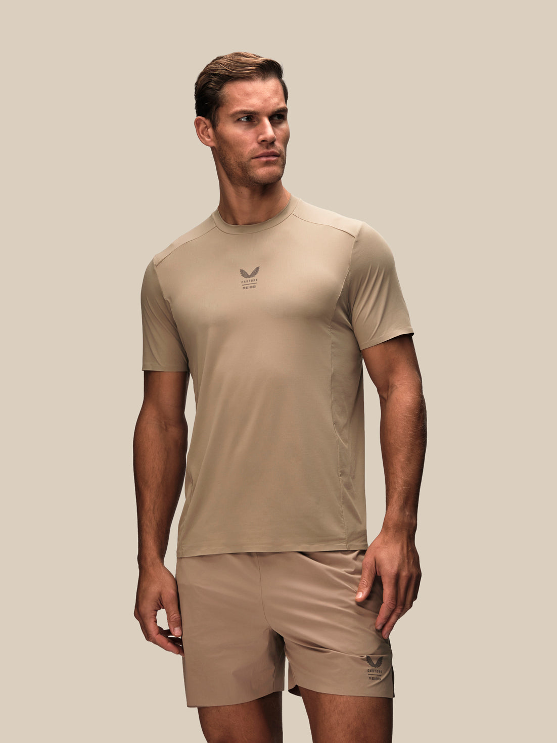 A man wearing a beige athletic short-sleeve t-shirt and matching shorts, standing against a solid beige background.
