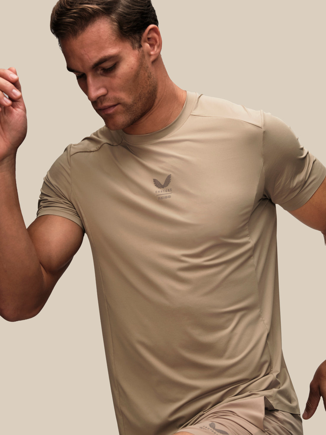 A man wearing a beige athletic short-sleeve t-shirt with a dark gray logo on the left chest, posed against a solid beige background.