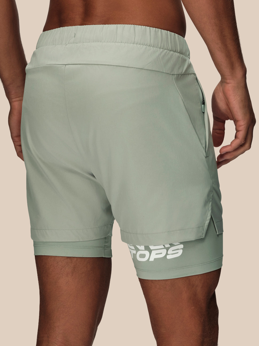 A close-up back view of a person wearing sage green athletic shorts with a white logo near the hem, set against a solid beige background.