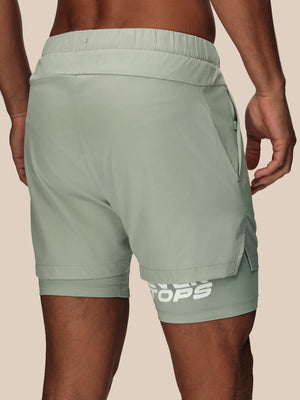 A close-up back view of a person wearing sage green athletic shorts with a white logo near the hem, set against a solid beige background.