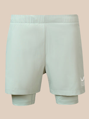A pair of light sage green athletic shorts shown from the back, featuring a built-in compression liner at the hem with a white 'STOP' logo on the left leg, set against a solid beige background.