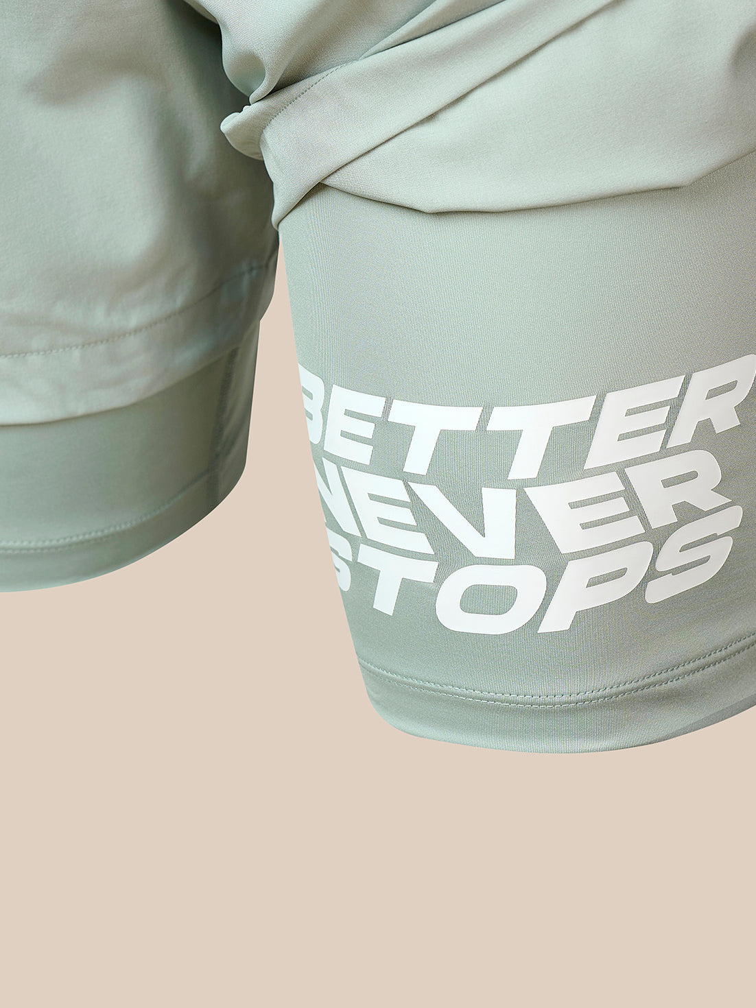Close-up of sage green athletic shorts with a large white graphic text reading 'BETTER NEVER STOPS' on the thigh, set against a solid pale beige background.