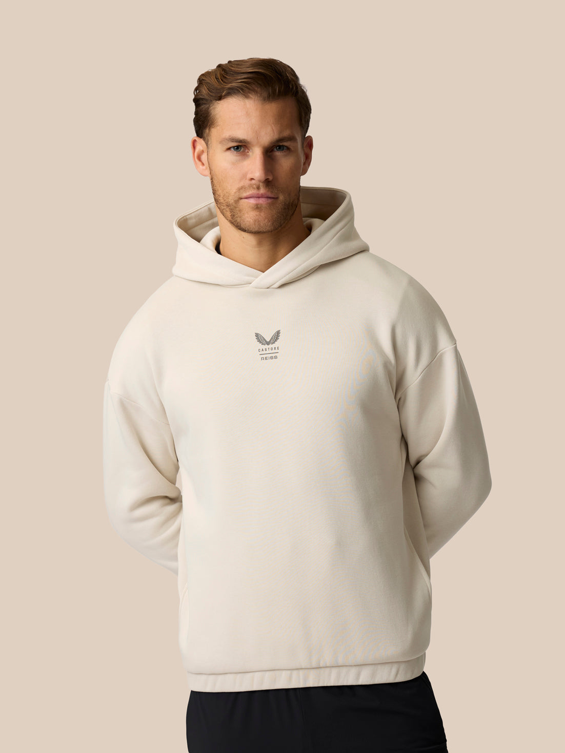 A man wearing a cream-colored textured hoodie with a small gray winged logo on the left chest, set against a solid beige background.