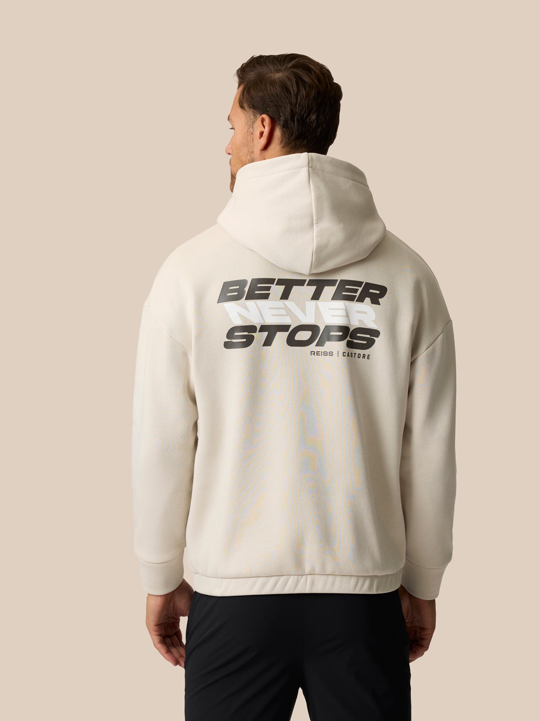 A man viewed from the back wearing an off-white hoodie with a large black graphic reading 'BETTER STOPS' on the back, paired with black pants, against a solid beige background.