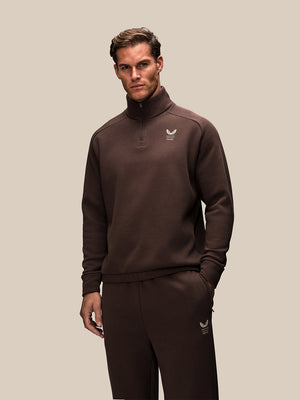A man modeling a dark brown quarter-zip sweatshirt and matching sweatpants set with a small white winged logo on the chest and thigh, set against a solid beige background.