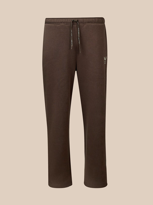 A pair of dark brown sweatpants with a drawstring waist and a small white embroidered logo on the left thigh, shown against a solid beige background.