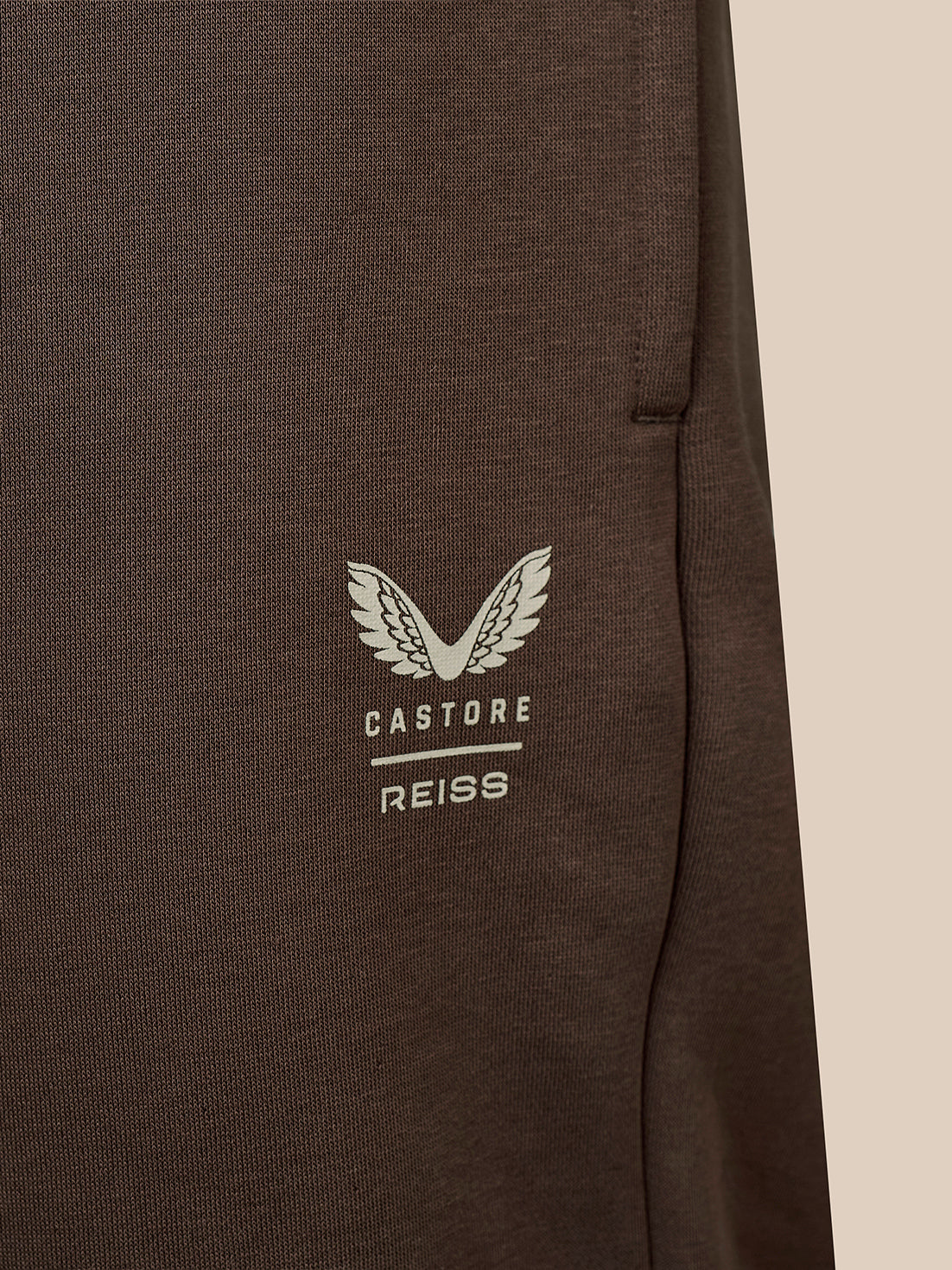 Close-up of a dark brown textured fabric garment featuring a white embroidered wing logo and the text 'CASTORE REISS' on the chest.