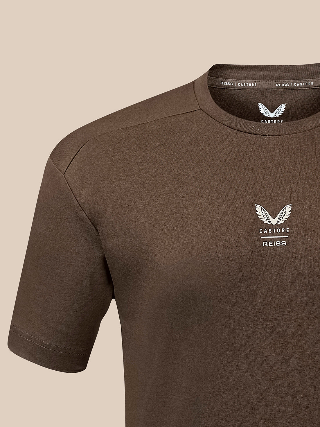 A close-up of a brown short-sleeve t-shirt with a white winged logo and the text 'CASTORE REISS' on the left chest, set against a solid beige background.