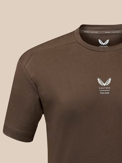 A close-up of a brown short-sleeve t-shirt with a white winged logo and the text 'CASTORE REISS' on the left chest, set against a solid beige background.