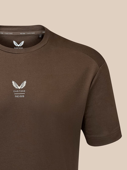 A close-up of a dark brown short-sleeve t-shirt with a white embroidered winged logo and the text 'CASTORE REISS' on the left chest, set against a solid beige background.