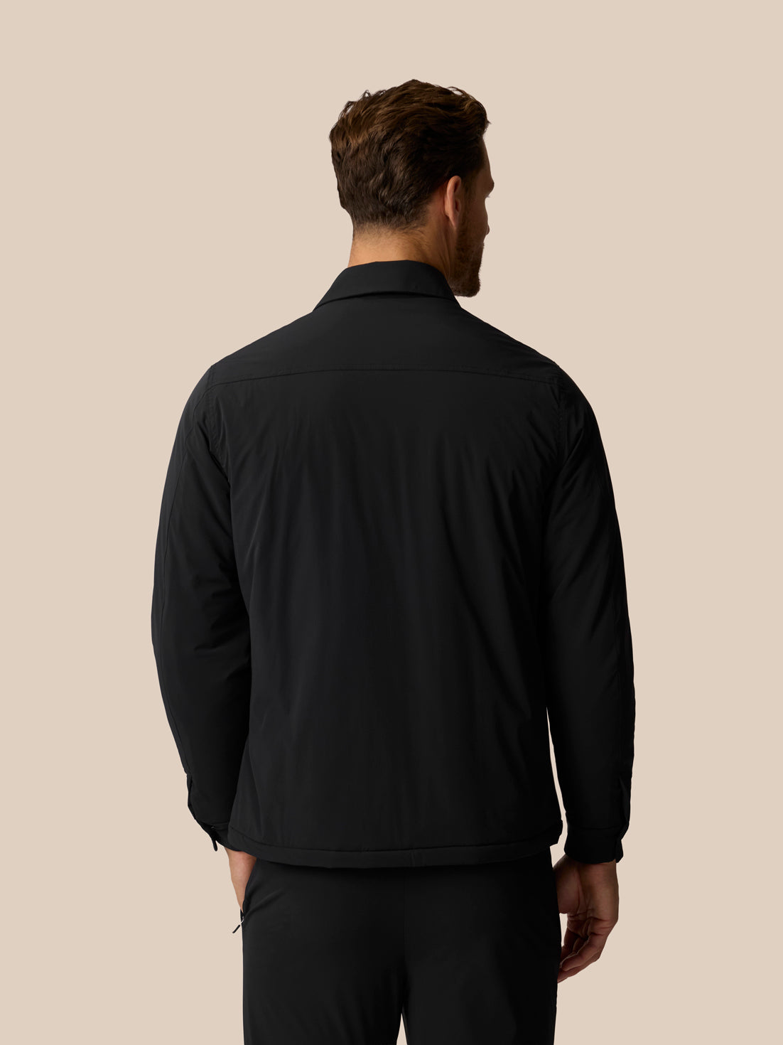 A man viewed from the back wearing a black long-sleeve collared jacket against a solid beige background.