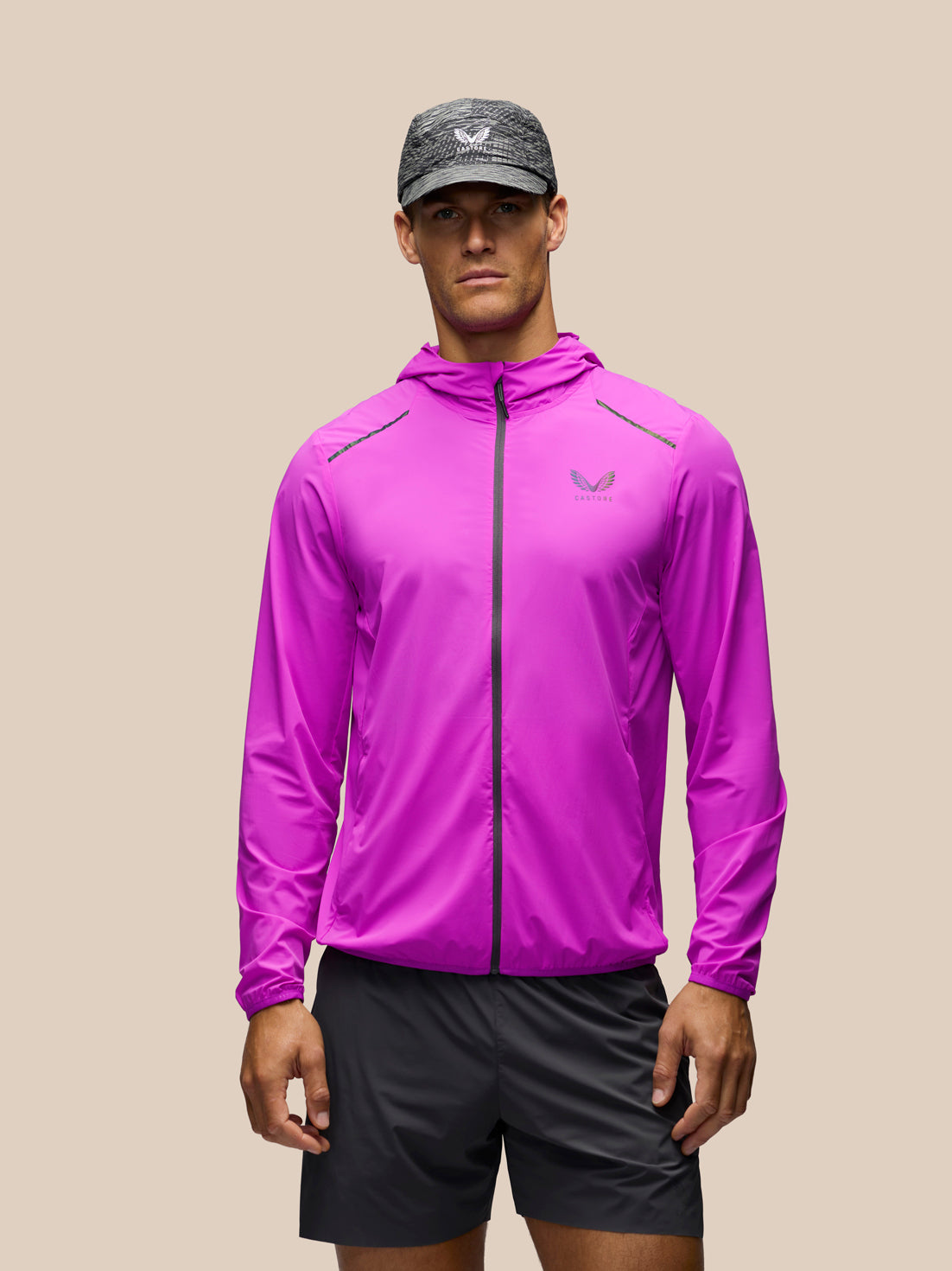 A man wearing a bright magenta-purple athletic windbreaker jacket with a black zipper, black running shorts, and a gray patterned cap, standing against a solid beige background.
