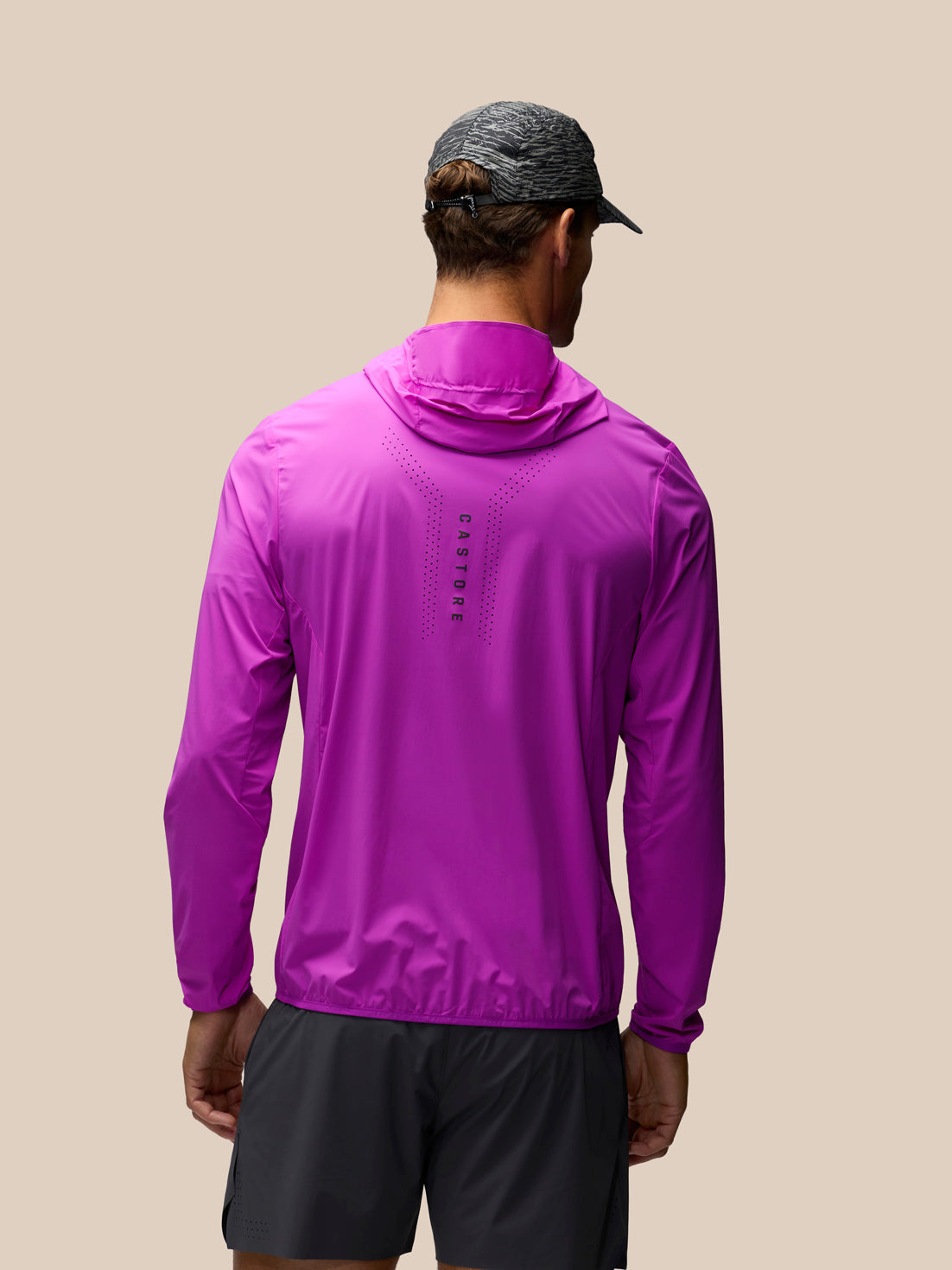 A man viewed from behind wearing a bright purple hooded long-sleeve athletic jacket and dark gray running shorts against a solid beige background.
