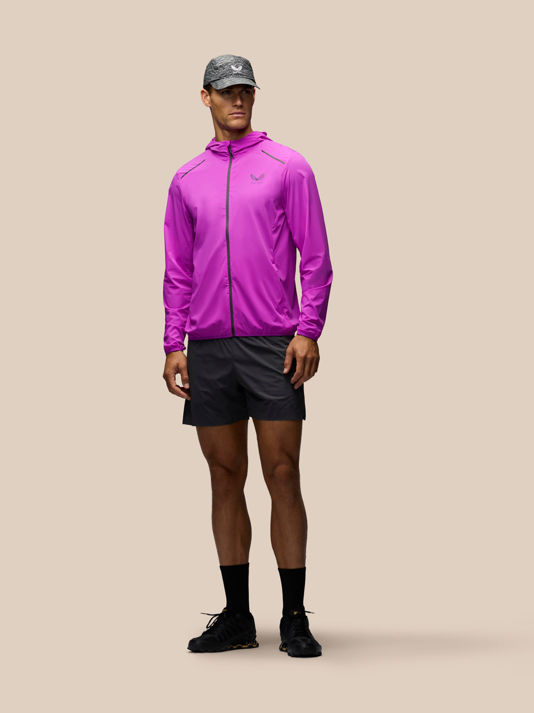 A male model wearing a bright purple athletic zip-up jacket, dark gray running shorts, a gray cap, black socks, and black running shoes, standing against a solid beige background.