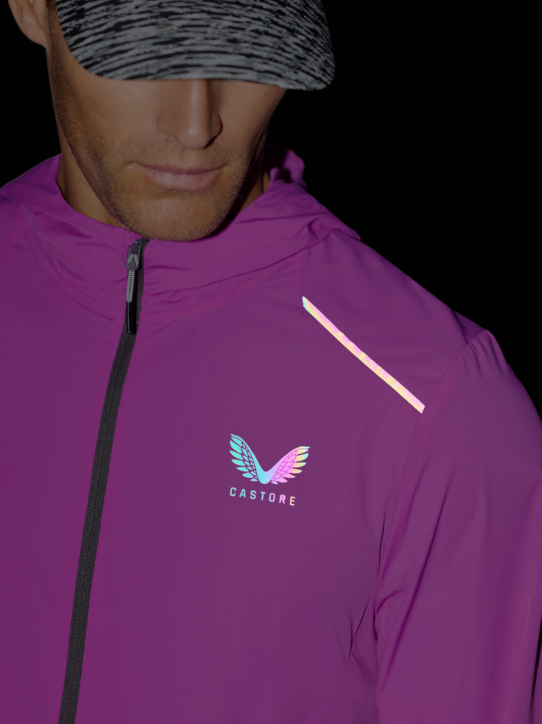 A close-up of a man wearing a purple Castore athletic jacket with a reflective diagonal stripe on the shoulder and a colorful winged logo on the chest, set against a dark background.