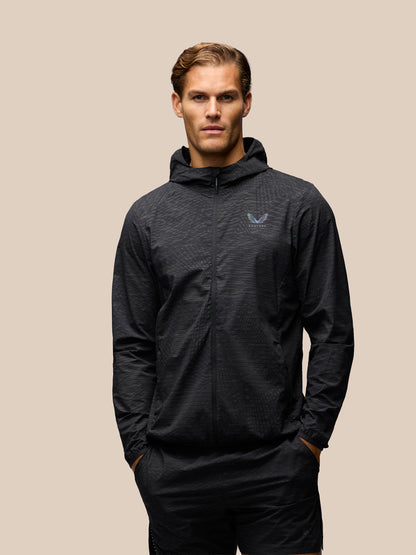 A man wearing a dark gray hooded zip-up jacket with a subtle textured pattern and a small blue logo on the left chest, posed against a solid beige background.