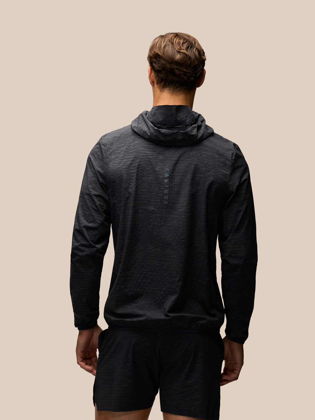 A man viewed from behind wearing a dark charcoal hooded long-sleeve athletic top and matching shorts against a solid beige background.