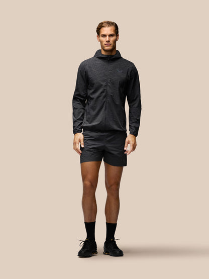 A male model stands against a solid beige studio background wearing a dark gray hooded long-sleeve athletic top, matching dark gray shorts, black socks, and black running shoes.