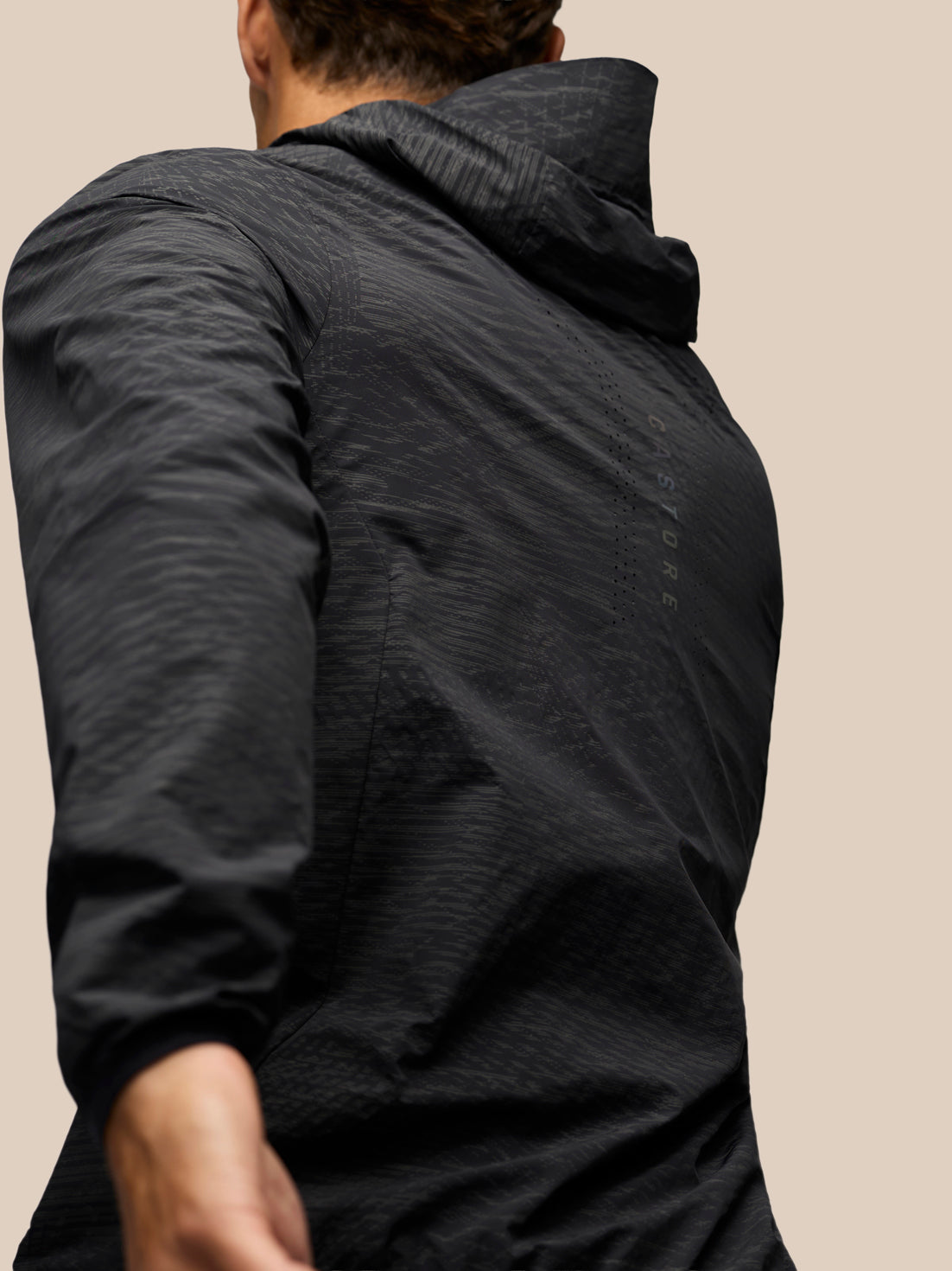 A close-up back view of a person wearing a dark charcoal gray hooded jacket against a solid beige background.