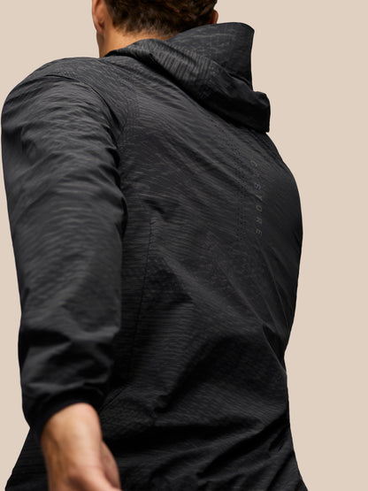 A close-up back view of a person wearing a dark charcoal gray hooded jacket against a solid beige background.