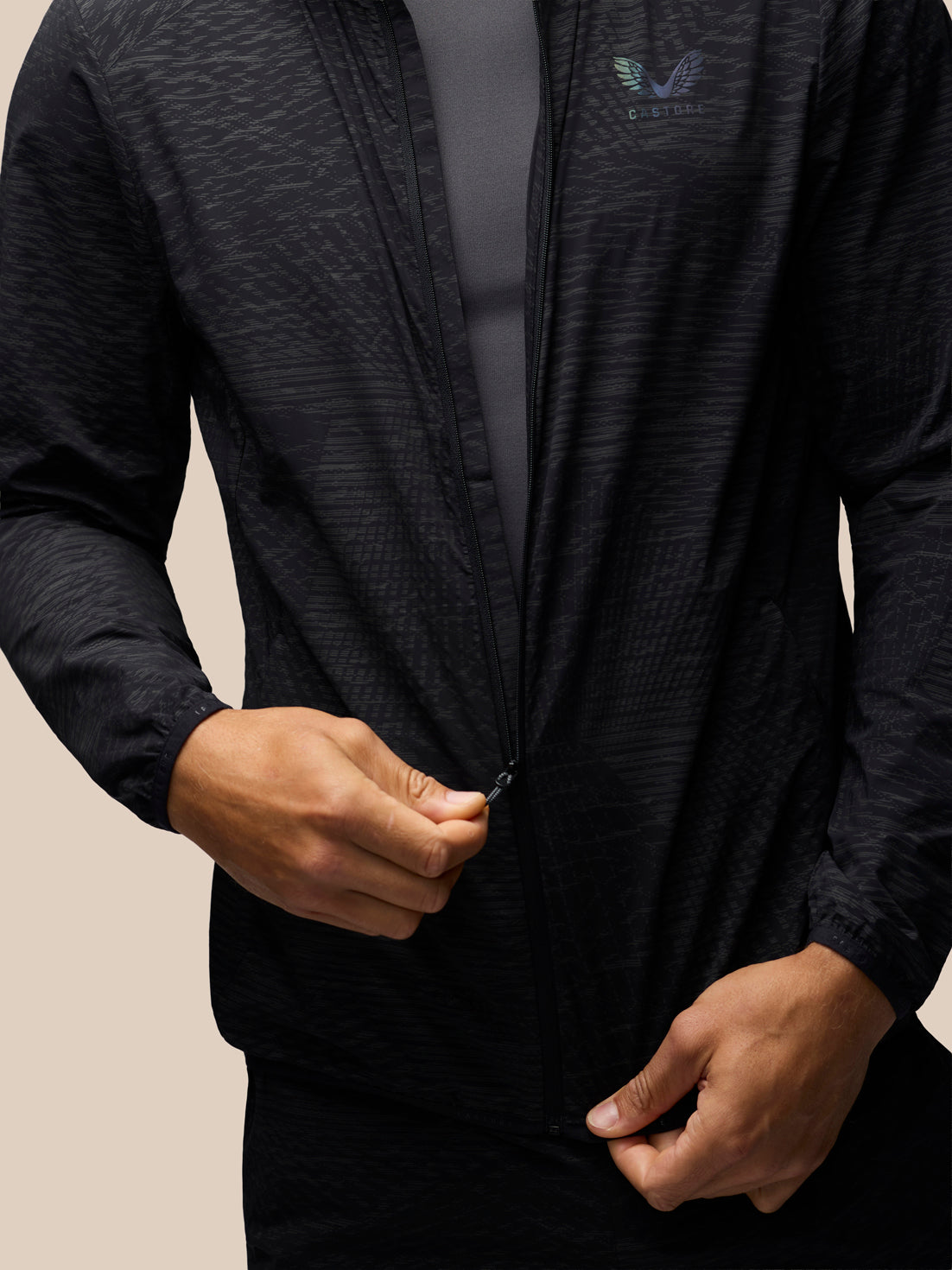 A close-up of a person wearing a black textured zip-up athletic jacket over a dark gray shirt, with the person's hands adjusting the zipper. The jacket features a subtle winged logo on the left chest.