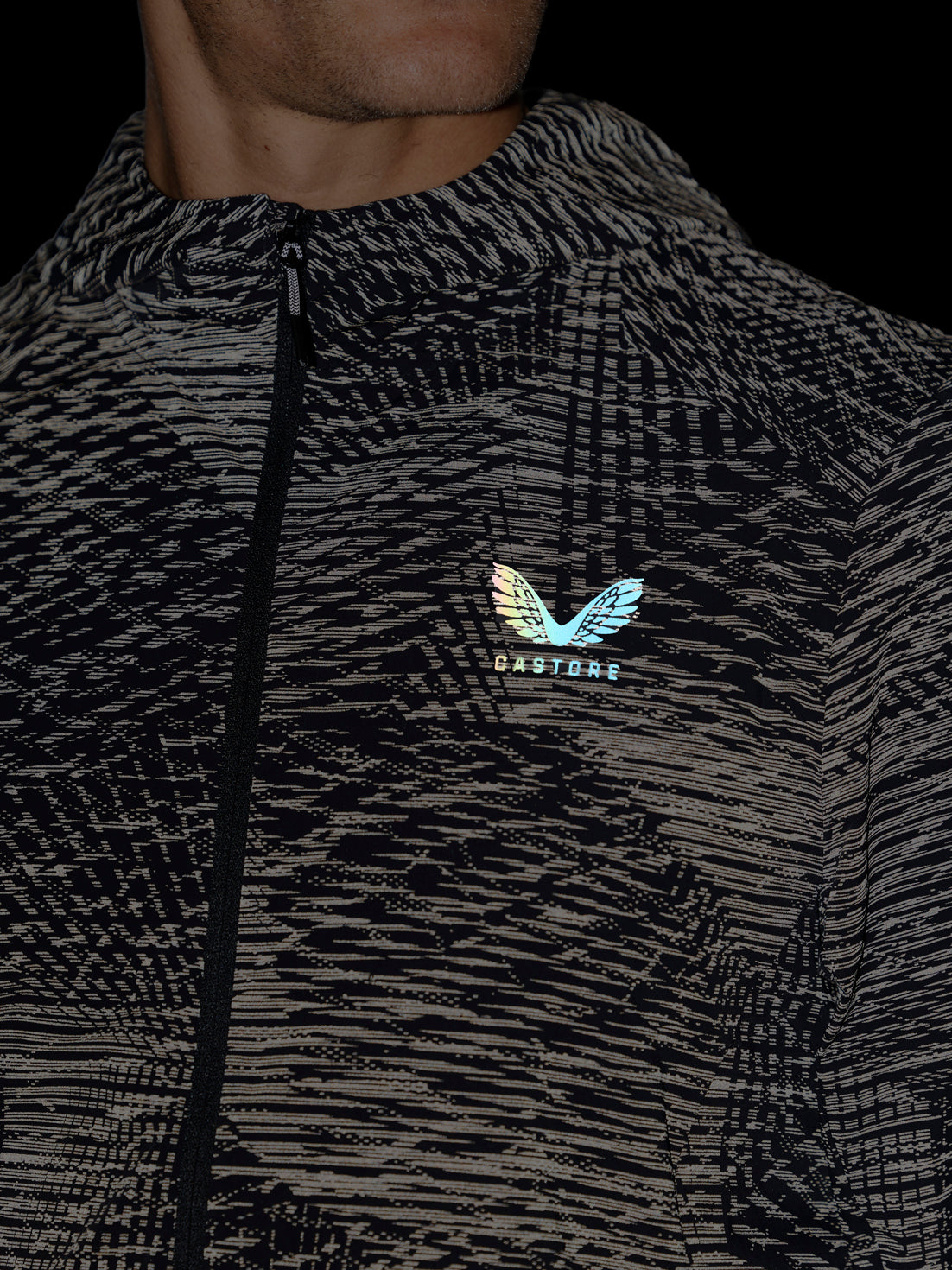 Close-up of a person wearing a black and gray patterned zip-up jacket with a small iridescent wing logo and the word 'CASTORE' on the chest, against a dark background.