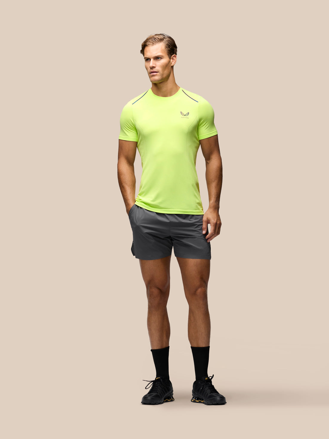 A male model stands against a solid beige background wearing a bright neon yellow-green athletic t-shirt with a small logo on the left chest, dark gray athletic shorts, black socks, and black running shoes.