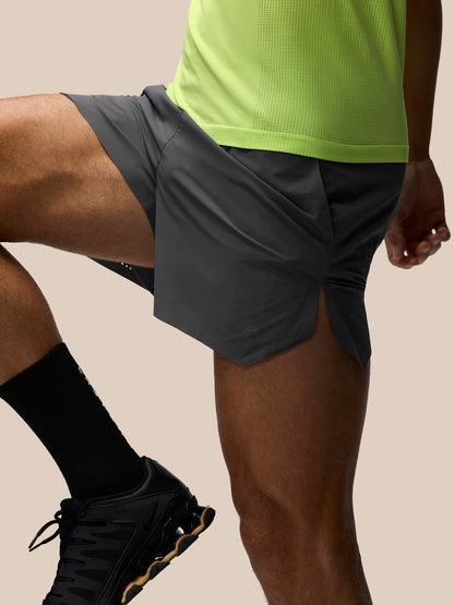 A close-up of a runner wearing dark gray athletic shorts with a side slit, paired with a neon yellow-green top, black socks, and black running shoes against a solid beige background.
