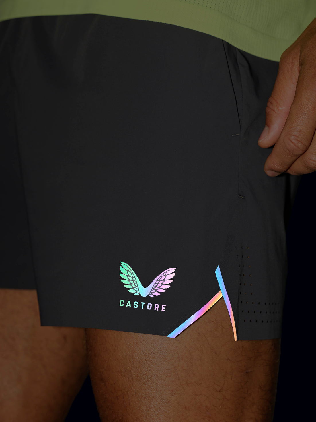 Close-up of a person wearing black Castore athletic shorts with a colorful wing logo and iridescent side accents, shown against dark clothing and skin.