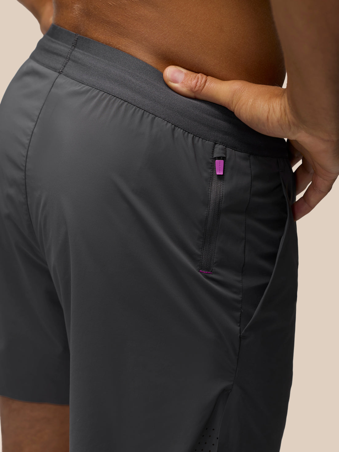 Close-up of a person wearing dark gray athletic shorts, showing the side pocket with a small purple tab and the waistband, against a plain beige background.