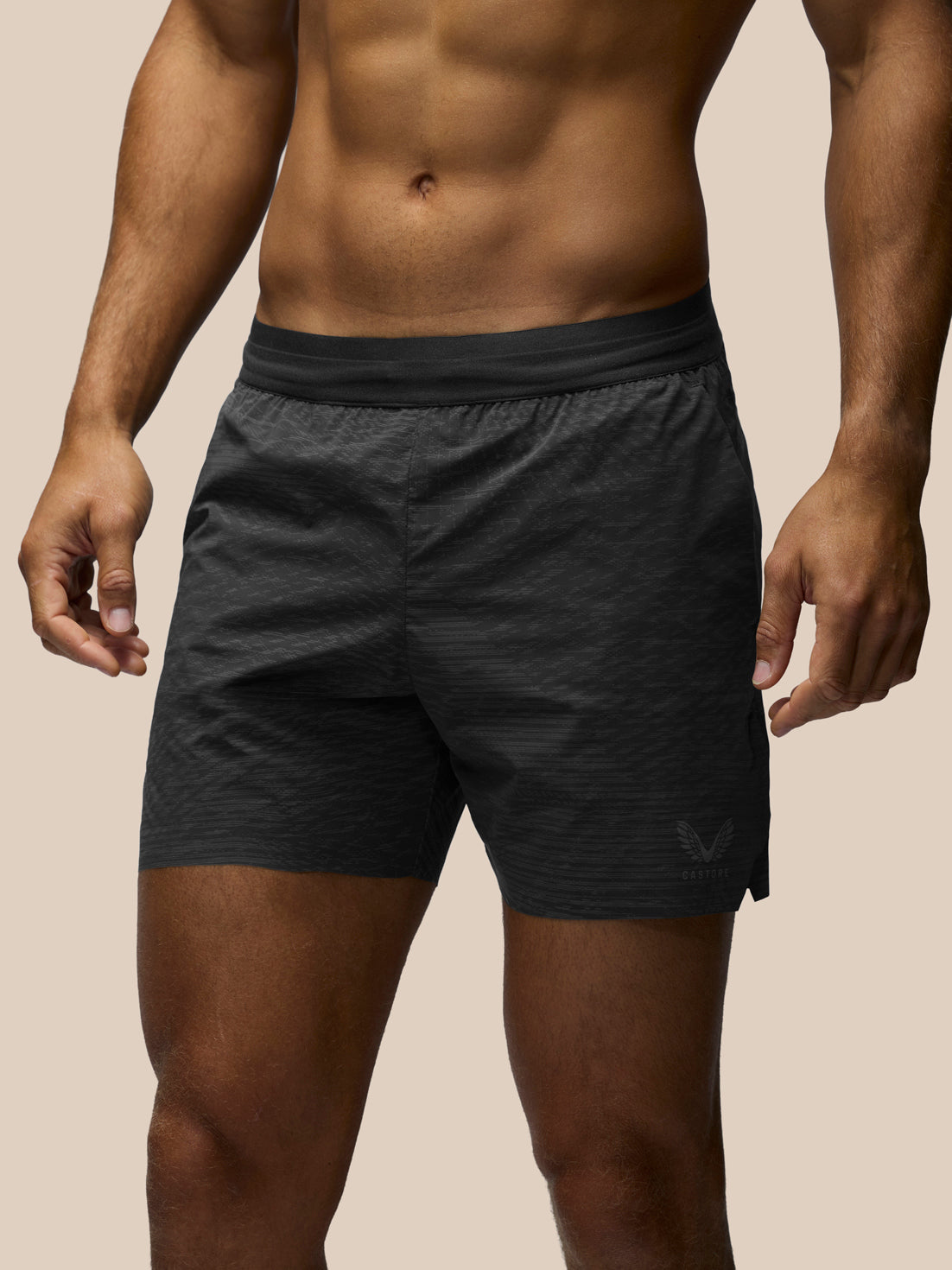 A close-up of a man's lower torso wearing dark charcoal gray athletic shorts with a subtle textured pattern and a small tonal logo on the lower right leg, set against a solid beige background.