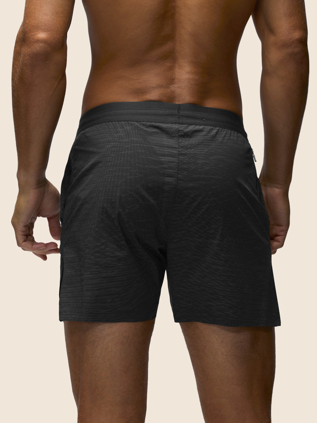 Rear view of a man wearing dark charcoal gray textured athletic shorts against a light beige background.