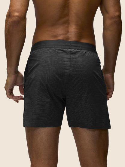 Rear view of a man wearing dark charcoal gray textured athletic shorts against a light beige background.