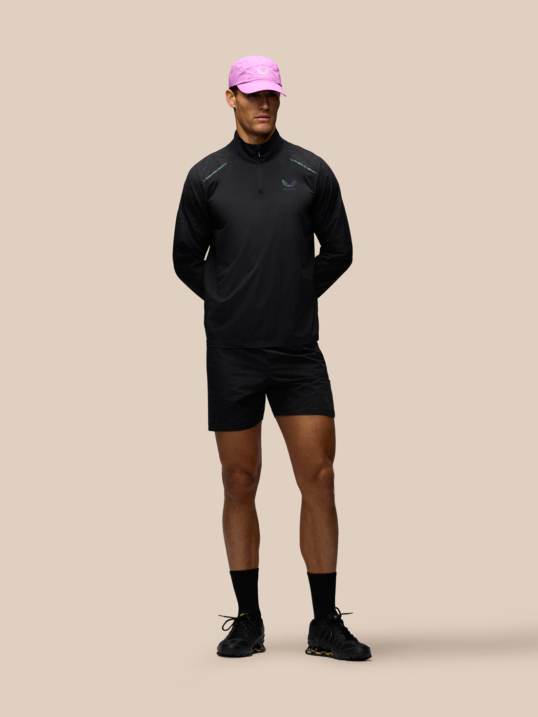 A male model stands against a solid beige background wearing a black athletic half-zip pullover, black shorts, black socks, and black running shoes, while wearing a bright pink baseball cap.