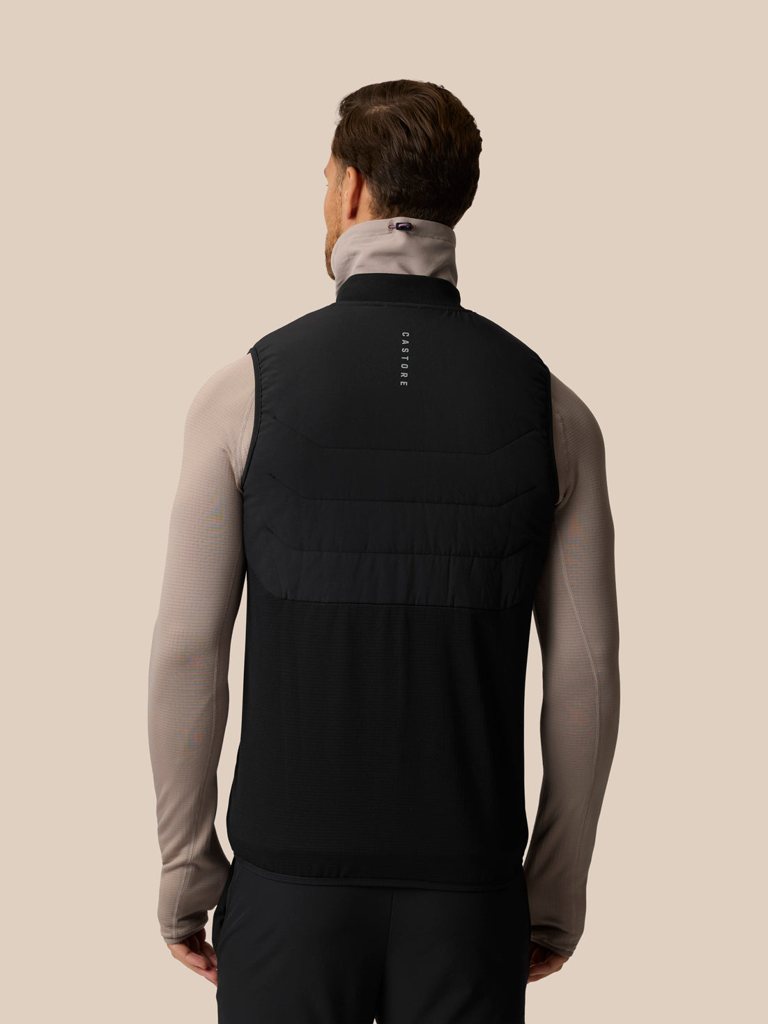 A man viewed from behind wearing a black sleeveless athletic vest over a light gray long-sleeve top, against a solid beige background.